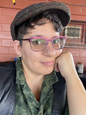 Shalimar smiling at the camera with their chin resting on their hand.  They are wearing a pink/purple glasses, a tweed patterned newsboy style  hat with red and blue speckles, and a black leather vest over a dark  green patterned shirt.  A curved barbell piercing is visible in one ear.