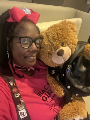 Bratty wearing glasses and a pink shirt with text, holding a teddy bear dressed in a leather vest.