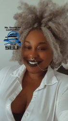 Bust photo of a Black femme presenting individual with light grey hair displayed in an afro like style smiling with black lipstick and one row of teeth showing. The individual is wearing a white button-up shirt with the top 3 buttons opened revealing cleaving and a small portion of a black undershirt. The Ms. SouthEast LeatherFest logo is displayed on the left of the photo near the individual's face.