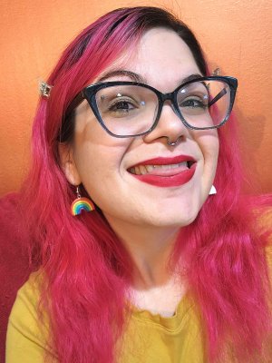 Photo of a smiling white woman with black-frame glasses, red lipstick, and long pink hair. She is in front of an orange wall, wearing a mustard-yellow shirt, and one rainbow earring is visible.