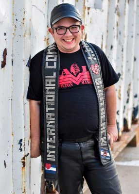 AJ stands smiling at the camera.  He's wearing a leather cap, dark rim glasses, a black T-shirt  and black leather jeans.  Draped over his neck is a title sash that reads International Community Bootblack  with a Bootblack pride flag.