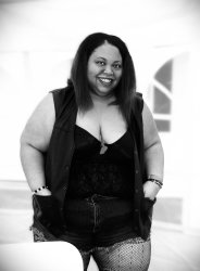 black and white photo with person standing with white tent background wearing shorts and leather vest open with shirt and center ribbon under vest, smiling face.