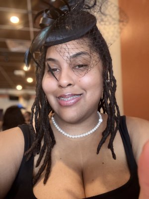 Woman seated smiling with black hat with veil on head with a pearl like necklace.