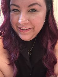 Picture of Bri’s smiling face with a lip piercing, two nostril piercings holding a gold chain draping across the top of their nose, a septum gold ring, and their long curly burgundy hair draping across their shoulders. Bri is wearing a mesh shirt under a black vest with their gold collar showing.