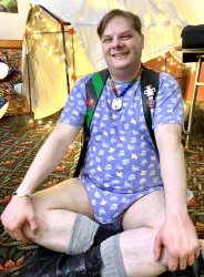 A young smiling person sitting on the floor cross legged wearing a blue onesie with toy cars and trucks on it,  wearing a leather vest and a rainbow chainmail collar with a lock. 
