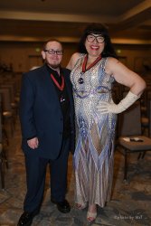 International couple 2024 standing together smiling. Master Retro Bella is on the right wearing a long dress, heels, forearm length gloves, and a medal around her neck. slave boy drake is on the left in a suit, dress shoes, and a medal around his neck.
