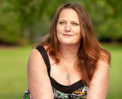 A headshot of Sara Blaze against a blurred green background. Sara has brown hair that flows down to her chest and is looking slightly up and to her right, squinting a little. She is wearing a silver polyamory symbol necklace and a tank top with comic references on it.