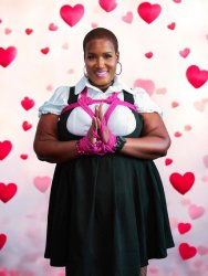 A smiling Black woman with a shaved head poses confidently in front of a pink and red heart-themed background. She wears a white puff-sleeve blouse under a black pinafore dress. Bright pink rope is styled in an artistic chest harness and wrapped around her wrists in a decorative bondage pattern. She has hoop earrings, subtle makeup with pink lipstick, and holds her hands together in a prayer-like gesture.