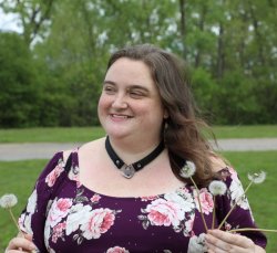 ShadowMe stands on a lawn with a dirt path and trees visible in the background. She has shoulder length brown hair.  She's wearing a plum-colored dress with  pink flowers and a black leather collar closed with a heart shaped silver padlock. She's holding several dandelions gone to seed.  She looks off to the left of the frame with a slight smile.