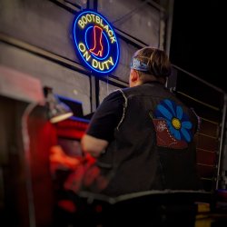 A person wearing a headband, wearing a jean vest with a blue daisy and a red combat boot on the back looking at a sign that reads Bootblack on duty