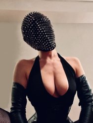 A half torso photo of Abundant Mischief. She is wearing a black sleeveless latex dress cut low past her chest and black gloves that go up to her biceps. Her face and hair are covered by a black fabric mask covered in silver studs.