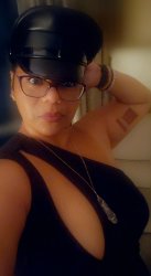 Master Lola Smiles posed with her torso at an angle and her face toward the camera with one hand behind her head. She wears a black master's cap, black-framed glasses, and a black dress with a teardrop cut out toward the shoulder closest to the frame. She also has gold earrings, a silver necklace, assorted bracelets on the hand behind her head, and a red tattoo on her upper arm.