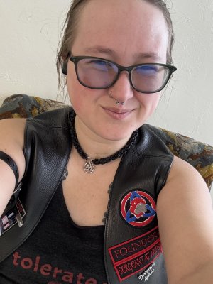 Luckyfyre wearing dark rimmed glasses, a black laced necklace with a silver Gordian knot charm, and two nose piercings.  They wear a black leather vest over a black tank top.  The vest includes several patches including ones that say  "Founder," "Sergeant at Arms," and "Designated Asshole."