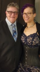 Two people standing lovingly together. Person on the left has short silver hair and is wearing dark rimmed eyeglasses,  black dress vest, blue striped collared shirt and a plaid black, grey, silver and blue tie. Person on the right has  dark purple hair and is wearing dark rimmed eyeglasses, a hoop septum nose ring, a thick silver slave collar, black  shawl over a navy blue lace dress and a black leather corset.
