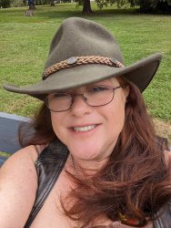 Patience Darling smiles at the camera.  She has auburn hair and is wearing dark rimmed glasses, a leather vest, and a gray felt rancher's hat.