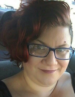 White woman in mid 40s with red curly hair and blue glasses