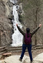 Elle stands on two large rocks with arms and legs outstretched, facing a distant waterfall with his back to the camera. His dark brown hair is in a braid. He is wearing a green jacket, a purple backpack, blue jeans, and hiking boots.