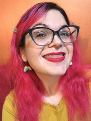 Photo of a smiling white woman with black-frame glasses, red lipstick, and long pink hair. She is in front of an orange wall, wearing a mustard-yellow shirt, and one rainbow earring is visible.