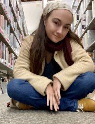 Femme adult with long brown hair in mustard yellow sneakers, jeans, a beige long sleeved top, and a beige bandana covering the top of their hair. They are sitting cross legged on the floor in between rows of books at a library.