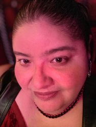 A close up face photo of Isabella Cross / slave maya. Her black hair is pulled back. She is wearing earrings, a black pearl necklace, a red top, and a black leather vest.