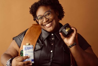 Daijah Flames is a black woman with black curly hair and dark frame glasses.  She wears a black button-up leather shirt with silver buttons and a brown leather vest.  She's smiling at the camera holding a can of Huberd's Leather Dressing in one hand and Obenauf's Heavy Duty Leather Preservative in the other.  She has a black bandanna wrapped around her right wrist.
