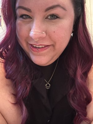 Picture of Bri’s smiling face with a lip piercing, two nostril piercings holding a gold chain draping across the top of their nose, a septum gold ring, and their long curly burgundy hair draping across their shoulders. Bri is wearing a mesh shirt under a black vest with their gold collar showing.